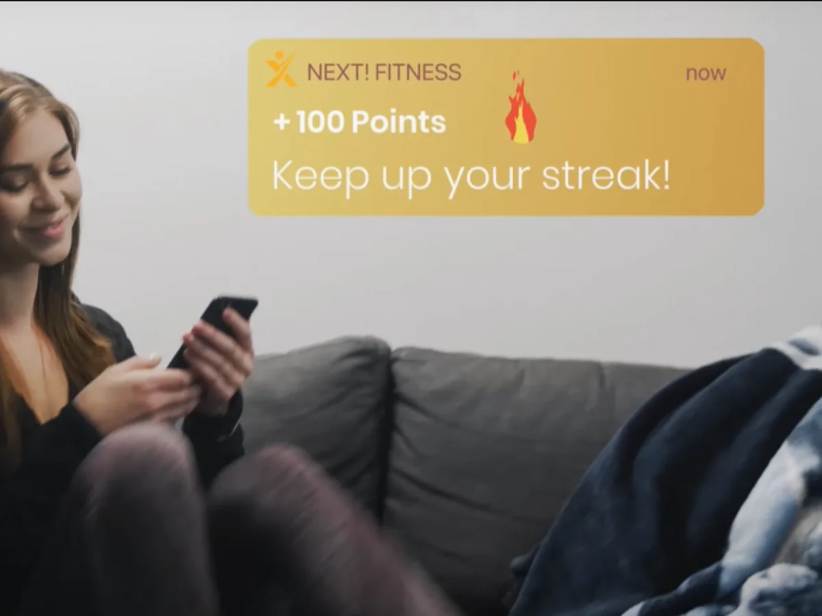 A member at home on the sofa reading a Next! Fitness notification: +100 points, keep up your streak